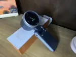 Handheld gimbal stabilizer resting on its box on a wooden table, with a round motor head facing the camera and a dark gray handle.